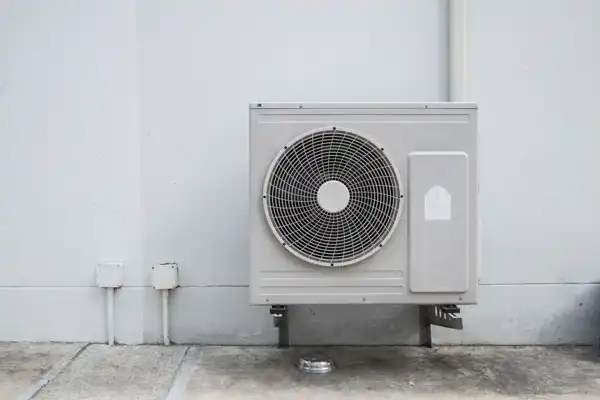 Wall-mounted outdoor air conditioning unit with a large fan, securely installed as a heat pump on a white wall above a concrete floor.