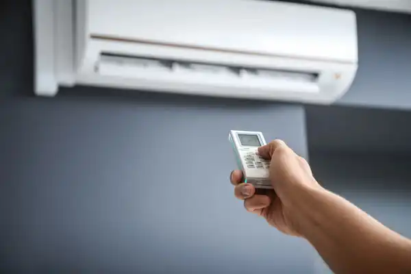 A hand holds a remote control pointing at a wall-mounted air conditioner against a blue-gray wall.