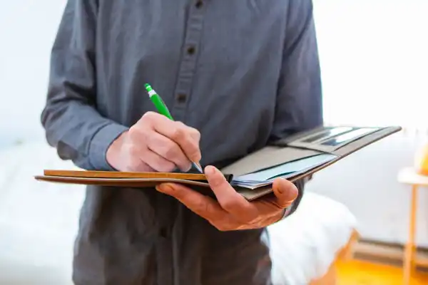 Person in a gray shirt writing with a green pencil on a notebook while standing.