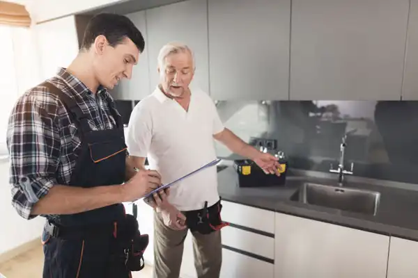 A plumber in overalls takes notes on a clipboard while an older man in a white shirt gestures towards the kitchen sink, with a toolbox visible on the counter.