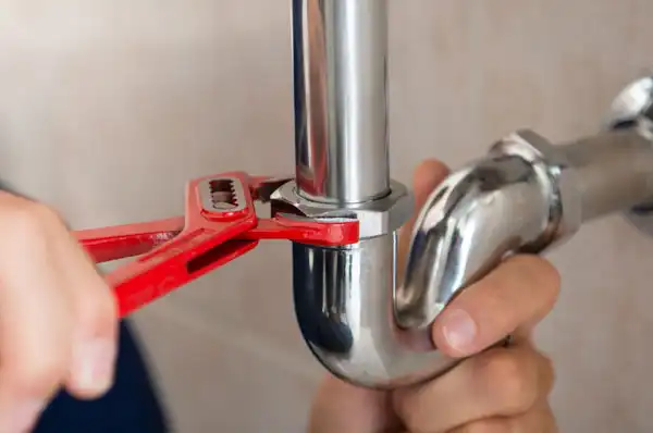 A person uses a red adjustable wrench to tighten a metal pipe fitting.