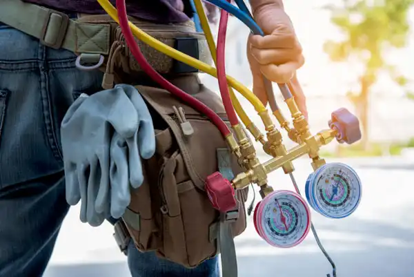 Person holding HVAC manifold gauges with hoses, wearing gloves attached to a tool bag.